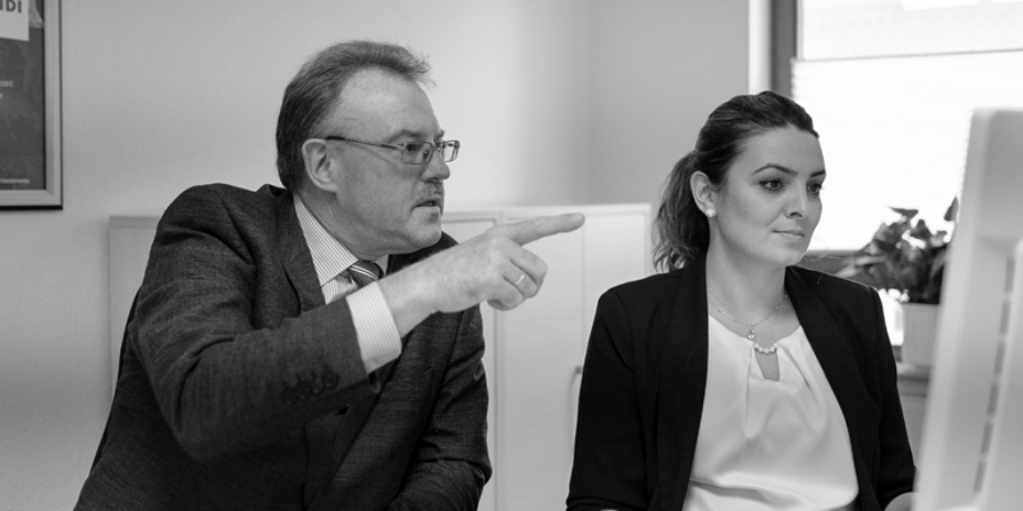 Thomas Sawade links und Mitarbeiterin rechts sitzen an einem PC, hinter Ihnen steht ein Regal und ein fenster. das bild ist schwarz-weiß.