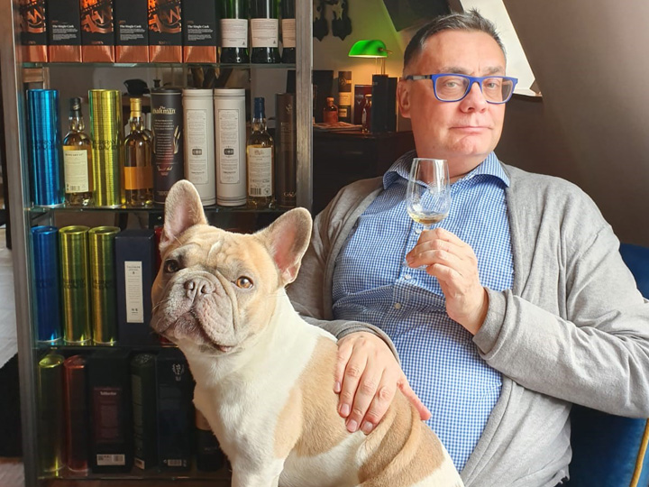 Wolf rainer Gust mit einem Glas in seiner rechten Hand. uaf ihm sitzt der Hund Boom un dhinter den Beiden ist ein Regal mit Flaschen