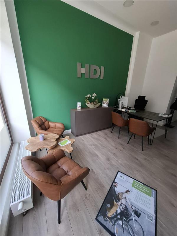 Das Büro wird gezeigt, links am Fenster stehen zwei braune Lederstühle zwischen den steht ein kleiner Holztisch. Hinten an der Wand hängt ein silberens HDI Logo und darunter steht ein graues Sideboard. Neben dem Sideboard steht ein Schreibtisch mit zwei braunen Lederstühlen davor und hinter dem Tisch steht noch ein Stuhl. Auf dem Tisch steht ein PC.