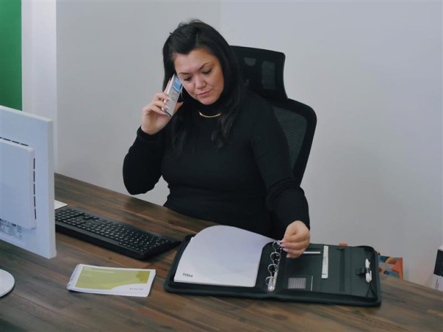 Frau Neubert sitzt am Schreibtisch und blättert in einem Ordner, indem viele Zettel drin sind. Außerdem telefoniert sie nebenbei, das sie sich das Telefon ans Ohr hält.