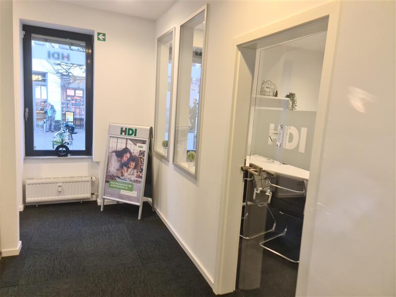 Man erkennt den Flur, rechts befindet sich ein Glastür, dahinter befindet sich ein Büro. Am Ende des Flurs ist links ein Fenster und rechts daneben steht ein HDI Aufsteller.