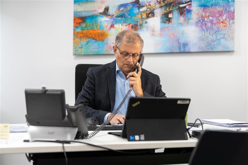 Der Agenturinhaber Alfred Hirt sitzt an einem Schreibtisch vor Laptop. Er hält in der rechten Hand ein Telefon an sein Ohr und tippt etwas in seine Tastatur, die vor ihm lieht. Hinter ihm an der Wand hängt ein buntes Bild.