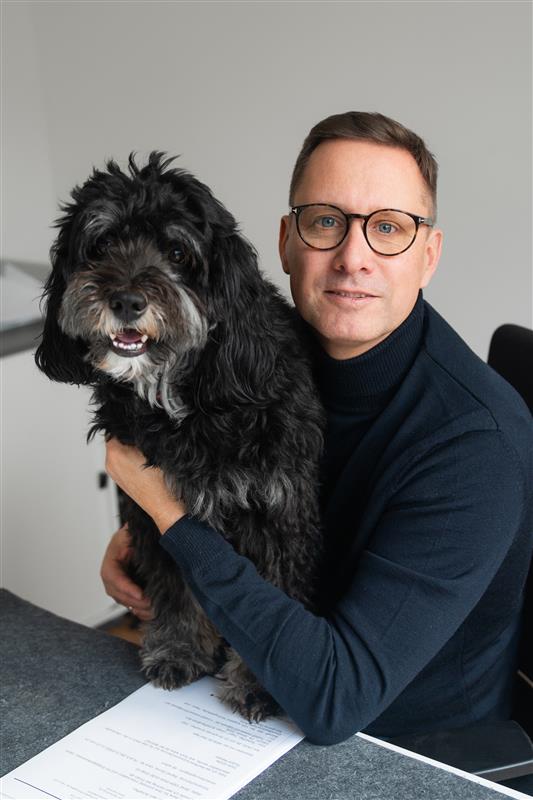 Scheffler sitzt am Schreibtsich und hält den Hund auf dem Schreibtisch fest, dabei guckt er in die Kamera.