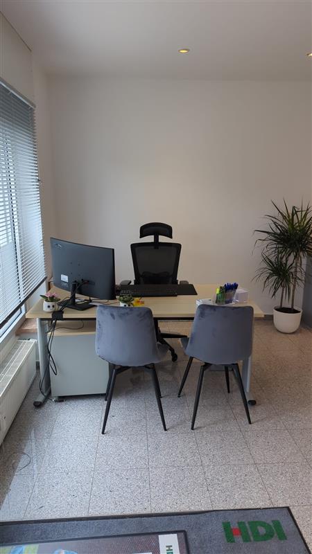 Im Büro steht ein Tisch an dem vorne zwei Stühle stehen und hinter dem Tisch ein weitere Stuhl. Auf dem tisch steht links ein PC und rechts neben dem Tisch steht ein Pflanze.
