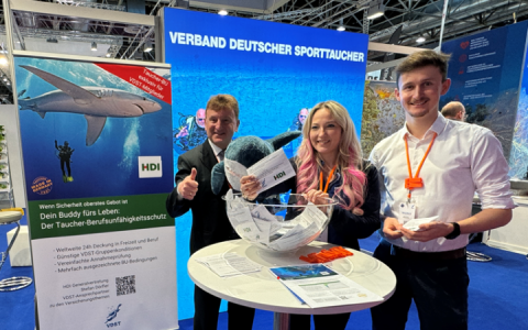 Dörfler und zwei Mitarbeiter befinden sich auf der Messe an dem Stand Verband Deutscher Sporttaucher und vor ihnen liegt ein Hai-Kuscheltier
