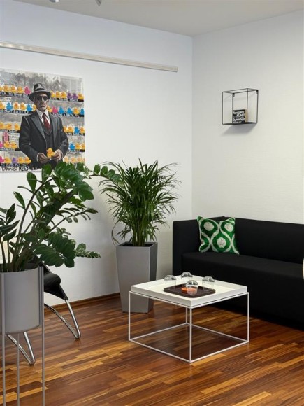 Rechts steht ein schwarzes Sofa, vor dem Sofa steht ein weißer Tisch. Links neben dem Sofa steht eine Pflanze. Links im Bild steht eine weitere Pflanze und hinter dieser Pflanze hängt ein Bild an der Wand. Auf dem Bild ist ein Mann zu erkennen.