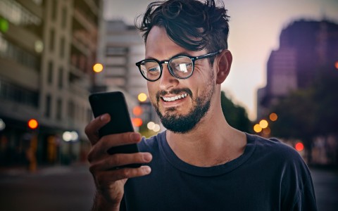Dunkelhaariger Mann mit Bart und schwarzer Hornbrille schaut lachend auf sein handy, im Hintergrund ist unscharf eine Straße mit Hochhäusern und Laternen zu sehen im Licht der untergehenden Sonne.