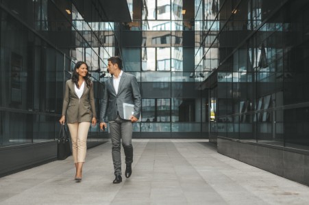 Zwei Geschäftsleute laufen selbstbewusst durch eine moderne Gebäudelandschaft und führen ein Gespräch. Die Szene vermittelt Professionalität und Mobilität im Arbeitsalltag.