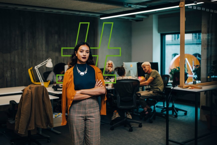 Eine Frau steht selbstbewusst in einem modernen Büro, im Hintergrund arbeiten weitere Personen; ein großes grafisches Symbol ist an der Wand sichtbar.
