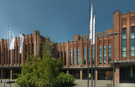 Außenaufnahme eines modernen Bürogebäudes der Regionaldirektion Köln bei Tageslicht mit roter Backsteinfassade und Flaggen vor dem Gebäude.