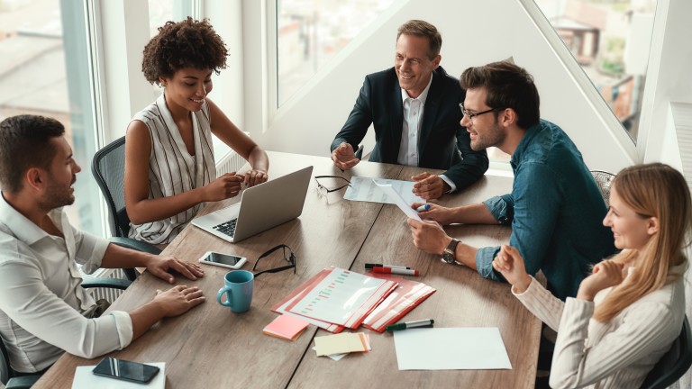 Mehrere Personen sitzen gemeinsam an einem Tisch und arbeiten an Laptops und Unterlagen in einer modernen Büroumgebung.