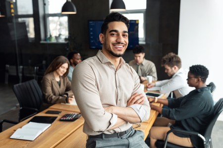 Ein Mann steht mit verschränkten Armen im Vordergrund eines modernen Büros, während im Hintergrund mehrere Kollegen an einem Tisch zusammenarbeiten und Laptops nutzen.