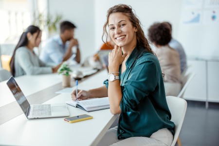 Eine Frau sitzt an einem Tisch und lächelt in die Kamera, während im Hintergrund weitere Kollegen gemeinsam an einem Meeting teilnehmen.