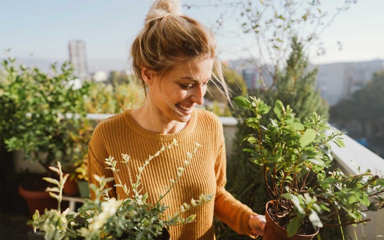 Frau mit hochgebundenem Haar in beigem Pullover auf Balkon oder Terrasse pflegt Pflanzen und hält einen Blumentopf