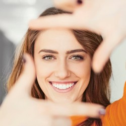 Frau lächelt in die Kamera und rahmt ihr Gesicht mit den Händen ein, unscharfer Hintergrund in Innenraum.
