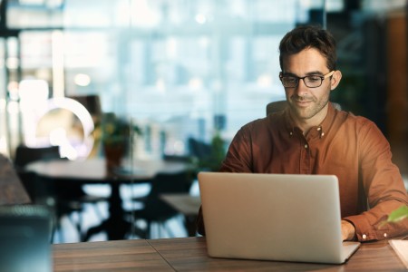 Mann mit kurzen Haaren, Brille und orangefarbenem Pullover sitzt am Schreibtisch und liest etwas am Laptop, im Hintergrund unscharf ein Büro.