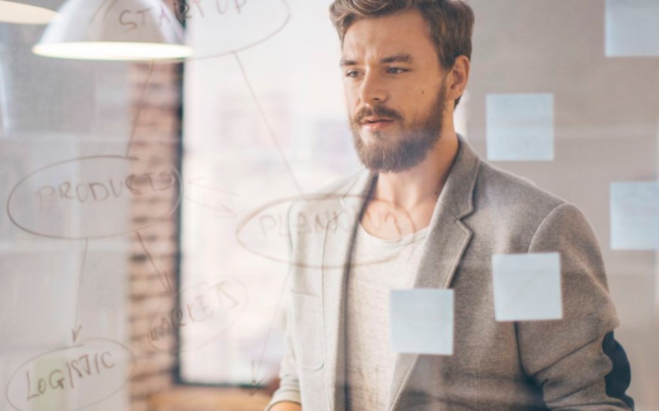 Blonder Mann mit Bart steht überlegend vor einem Ideen-Board in einem Büro, im Hintergrund eine Backsteinwand und ein Fenster