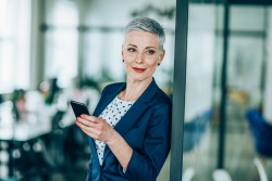 Businessfrau mit ganz kurzen grauen Haaren im Anzug lehnt an einer Bürotür und schaut lächelnd in die Ferne