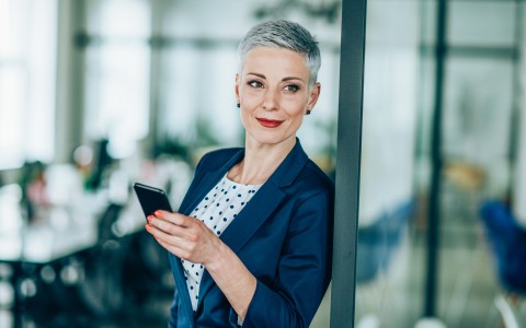Businessfrau mit ganz kurzen grauen Haaren im Anzug lehnt an einer Bürotür und schaut lächelnd in die Ferne