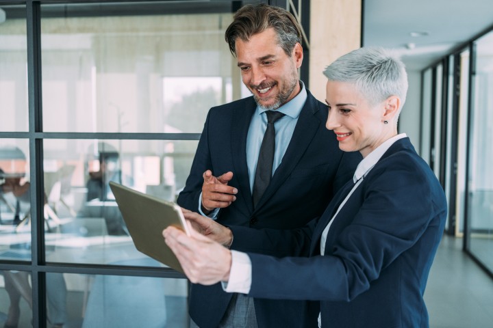 Zwei Businessleute - ein dunkelhaariger Mann und eine Frau mit ganz kurzen blonden Haaren - unterhalten sich in einem modernen Büroflur, während er ihr etwas auf dem Tablet zeigt