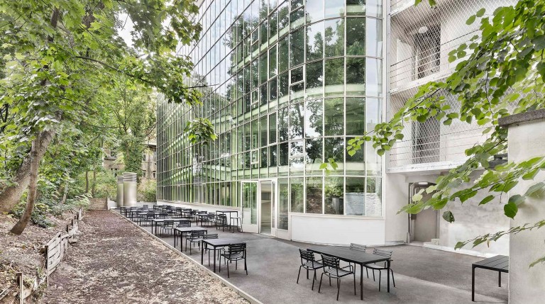 Modernes mehrstöckiges Bürogebäude aus Glasfronten umgeben vonhohen Bäumen, auf einem Vorplatz stehen mehrere graue Tosche mit Stühlen