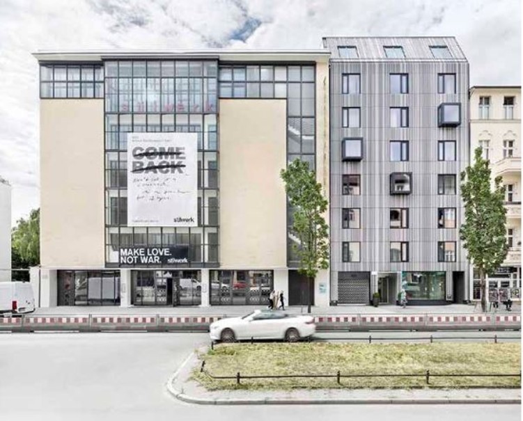 Blick auf eine moderne Häuserreihe mit Büros, Wohnungen, Geschäften, das direkt an einer Straße liegt,m davor steht ein weußes Auto