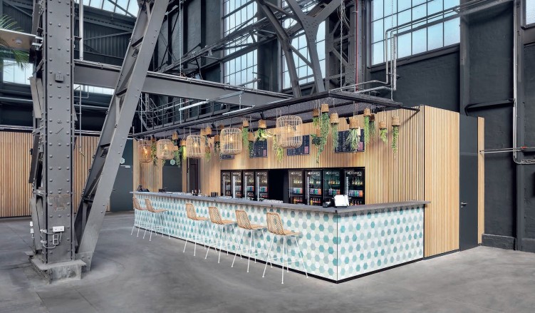 Große Halle - ähnlich einer Messehalle - mit schwarzen Stahlkonstruktionen,Blick auf eine holzverkleidete Bar mit mehreren Korblampen darüber