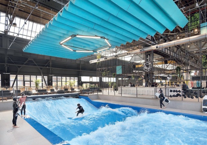 Riesige Halle - ähnlich einer Messehalle - mit einer Surfpool oder stehende Welle für Surfer in der Mitte, auf der einer Surfer surft und drei weitere am Rand um die Welle stehen