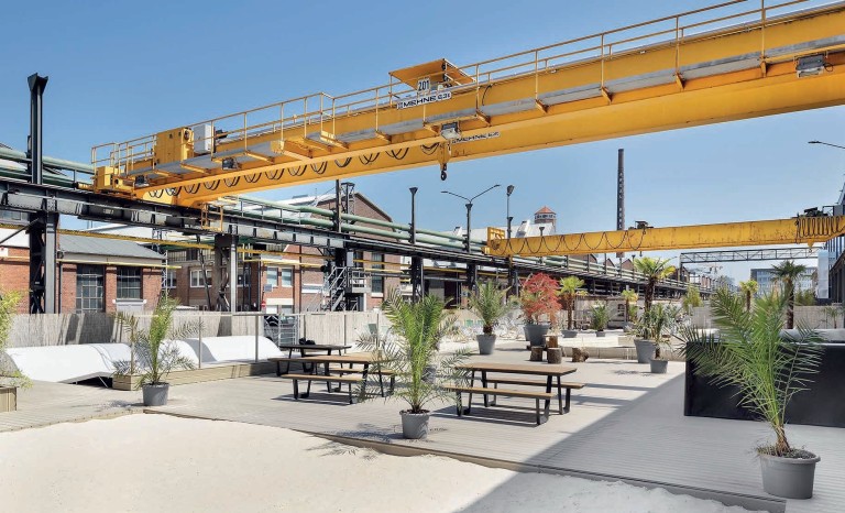 Eine moderne, industriell gestaltete Außenfläche mit Holzplanken, Palmen in Töpfen und Sitzbänken, die unter einer gelben Krananlage und Rohrleitungen verläuft.