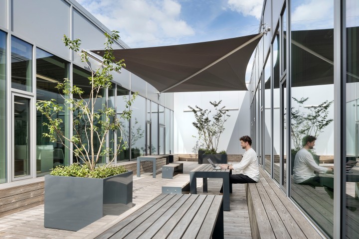 Mann sitzt in weißem Hemd auf einer modernen, begrünten Dachterrasse an einem Holztisch auf einer Holzbank und arbeitet an einem Laptop