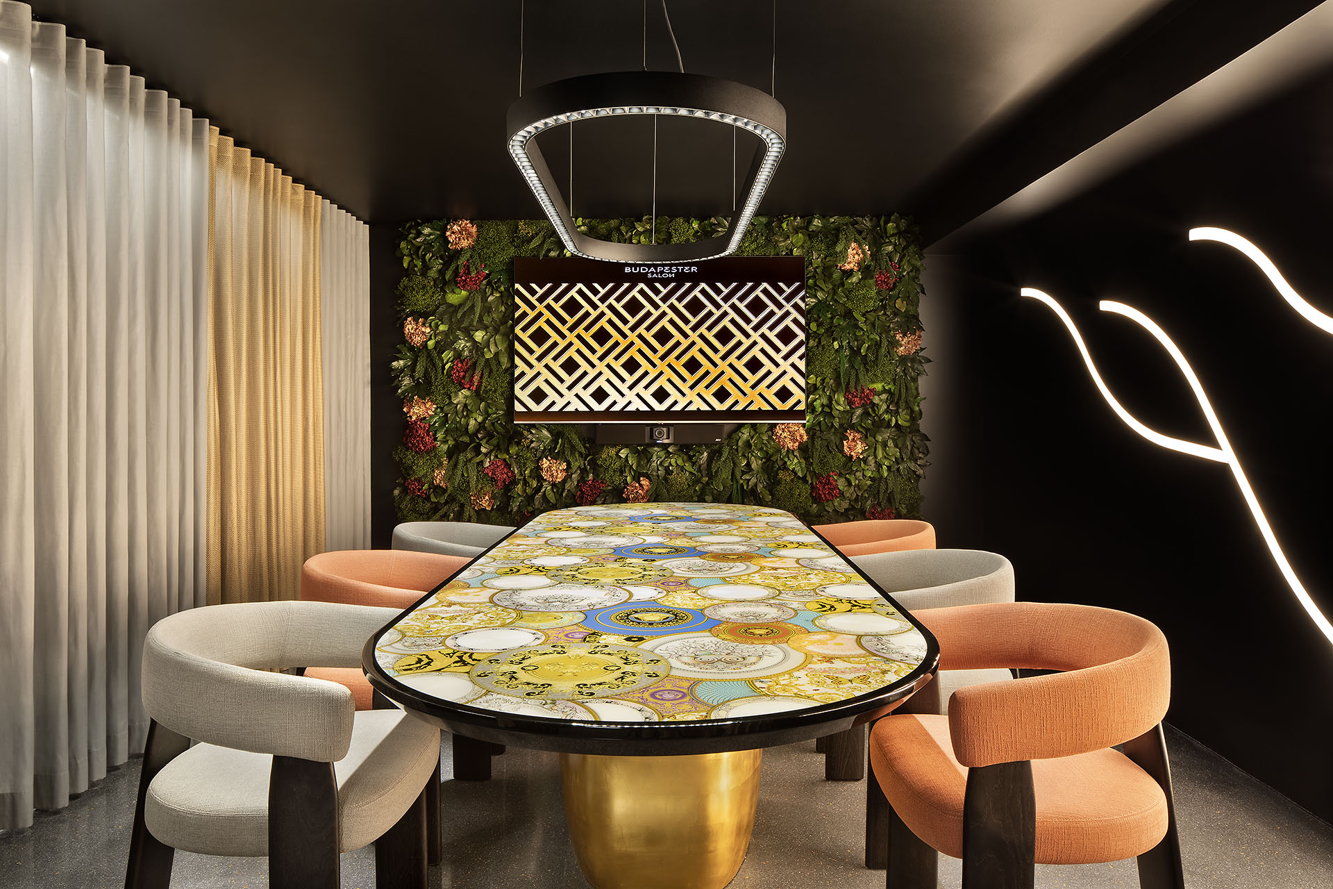 Innenansicht Budapester Salon mit einem großen Tisch mit großflächigem Mosaik auf der Platte, goldenem Fuß, darum Sessel in Naturtönen, die linke Wand besteht aus cremfarbenen Faltenvorhängen, die Kopfwand ist eine Blumentapete in dunklen Tönen und die Decke ist schwarzt