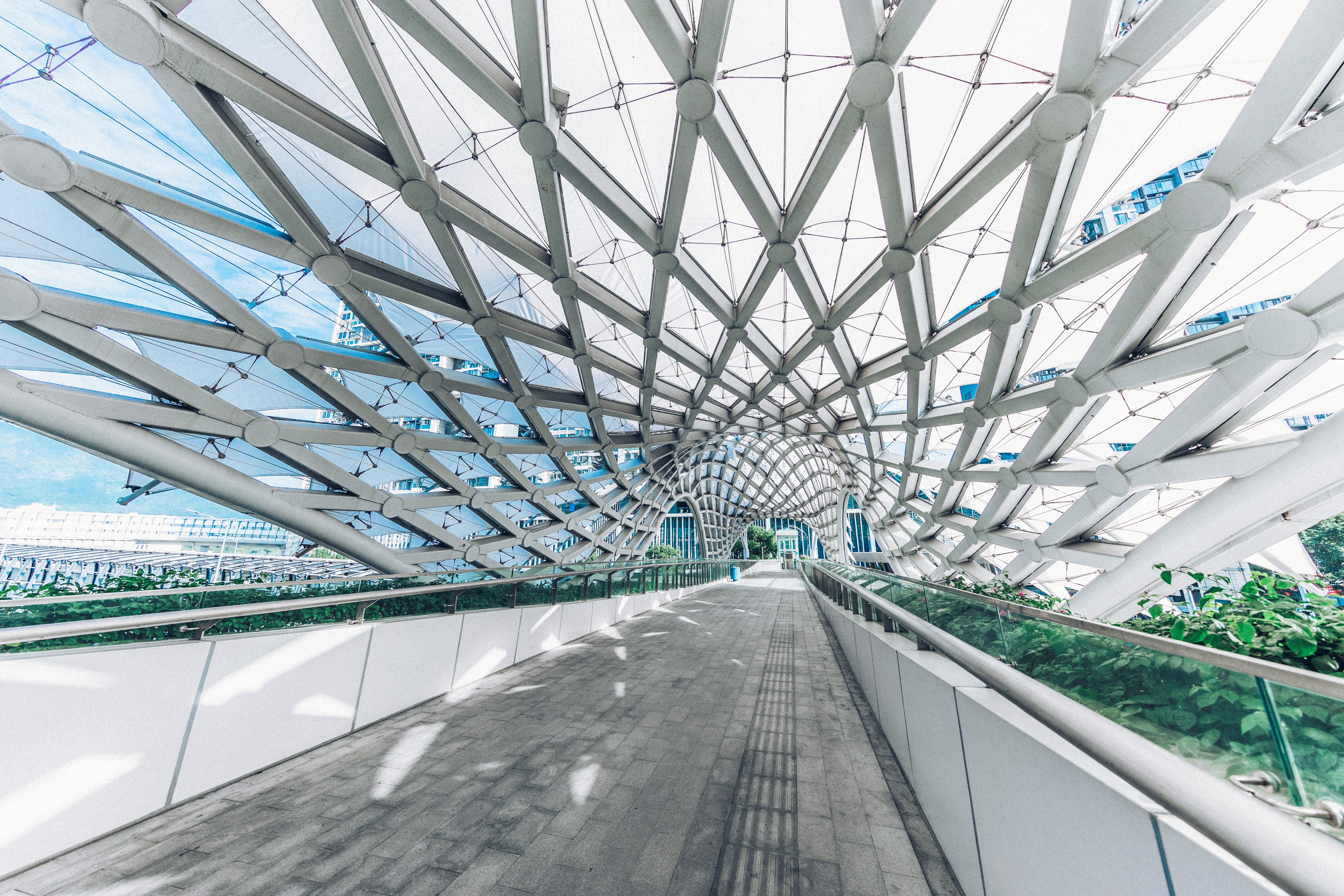 Innenansicht einer modernen, architektonischen Fußgängerbrücke mit einem auffälligen, wellenförmigen Dach aus weißen Stahlträgern und Netzen, das das Tageslicht filtert.