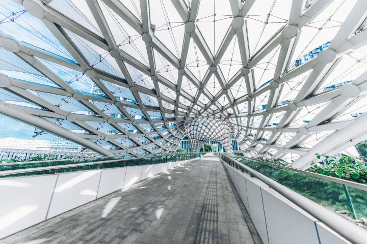 Innenansicht einer modernen, architektonischen Fußgängerbrücke mit einem auffälligen, wellenförmigen Dach aus weißen Stahlträgern und Netzen, das das Tageslicht filtert.