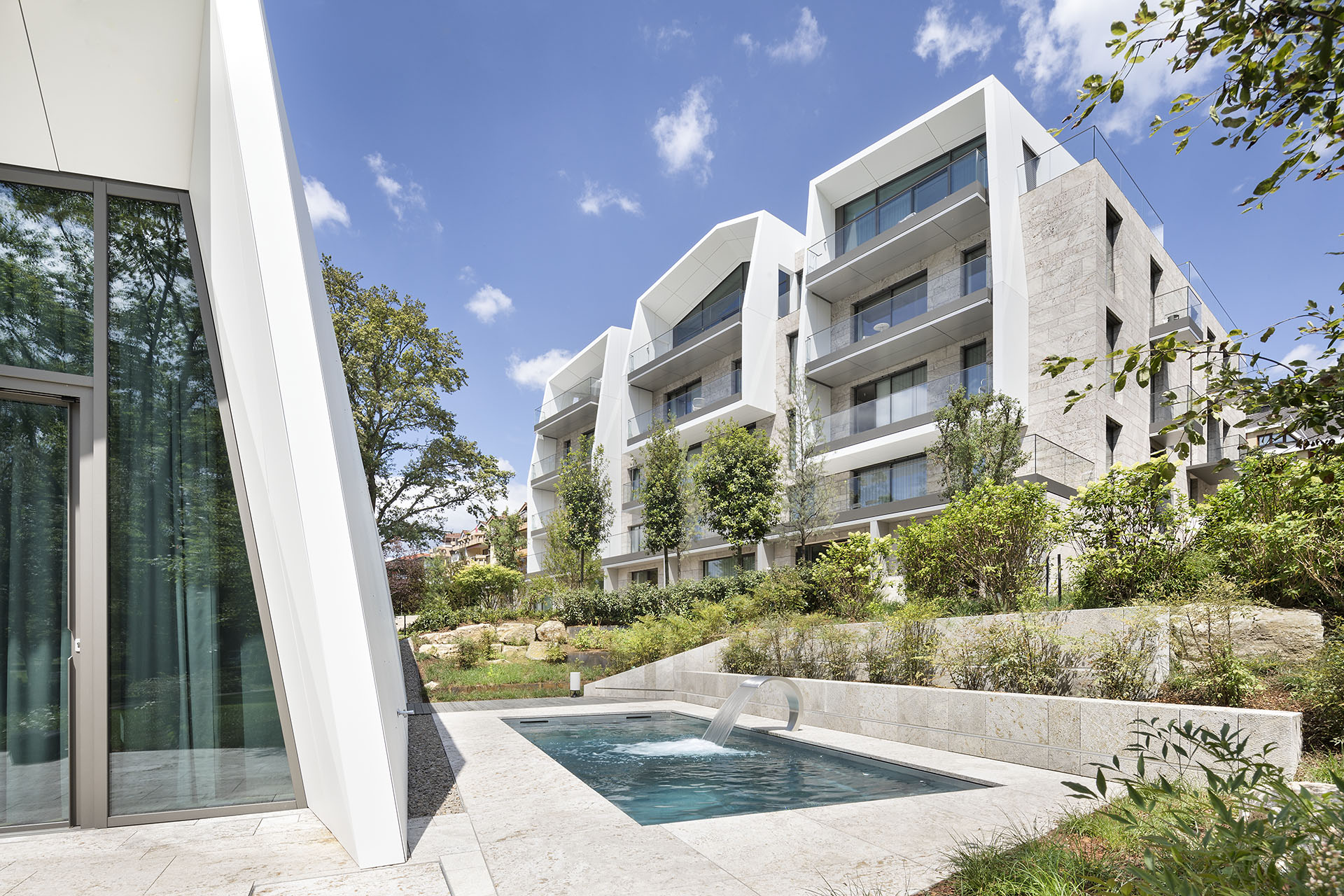 Moderne, zweigeschossige Luxus-Wohnblöcke mit Balkonen und einem Pool mit viel grün drumherum zwischen den Wohnblöcken im Sonnenlicht