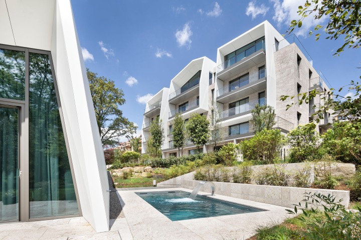 Moderne, zweigeschossige Luxus-Wohnblöcke mit Balkonen und einem Pool mit viel grün drumherum zwischen den Wohnblöcken im Sonnenlicht