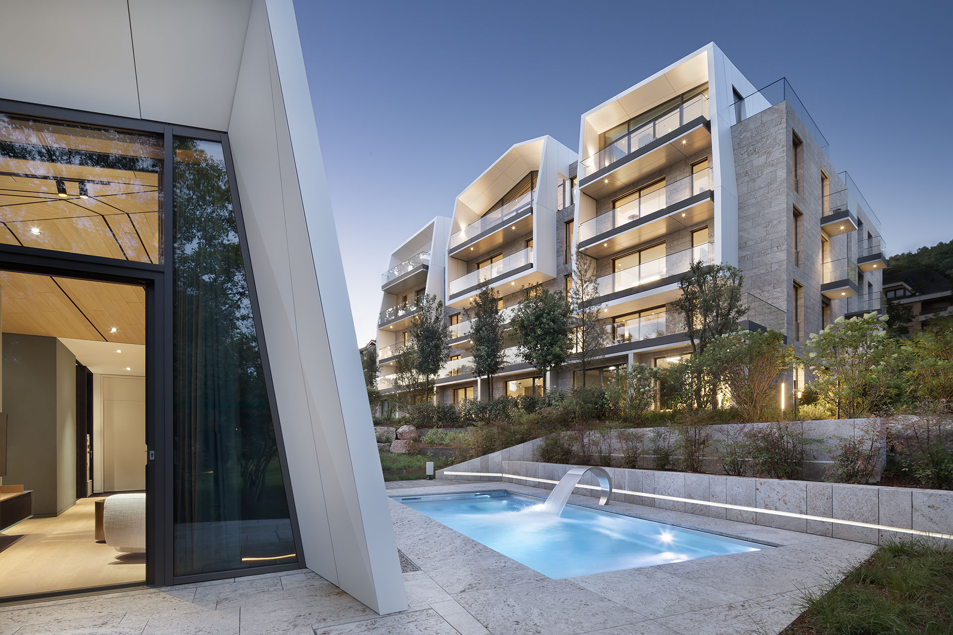 Moderne, zweigeschossige Luxus-Wohnblöcke mit Balkonen und einem beleuchteten Pool mit viel grün drumherum zwischen den Wohnblöcken im Abendlicht