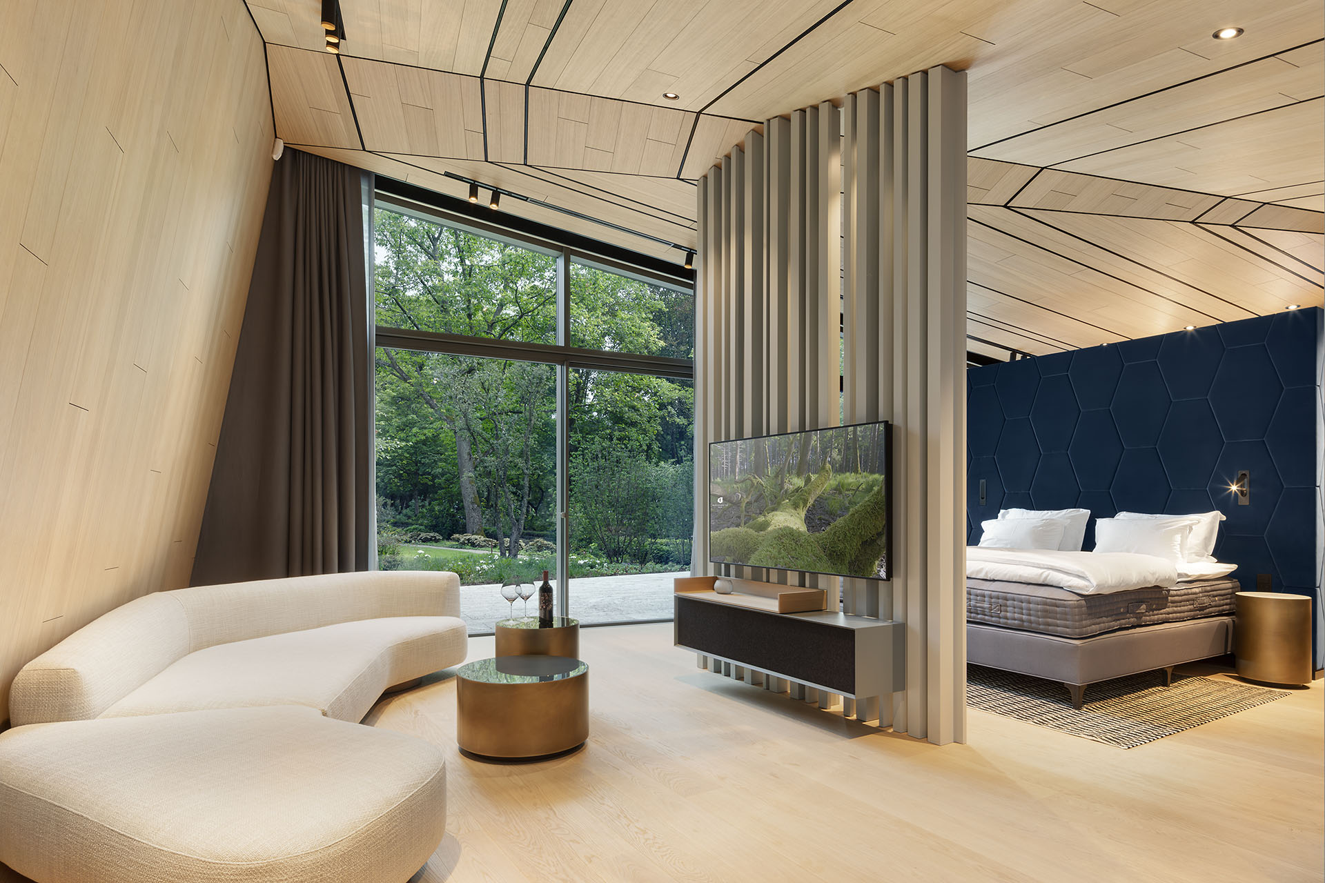 Modernes Hotelzimmer in hellen Beige- und Brauntönen, helles Holz an Wänden, Decke und Böden, cremefarbenes futuristisches Sofa, Flatscreen an einem Raumtrenner, Blick durch eine große Fenterfront in einen Garten