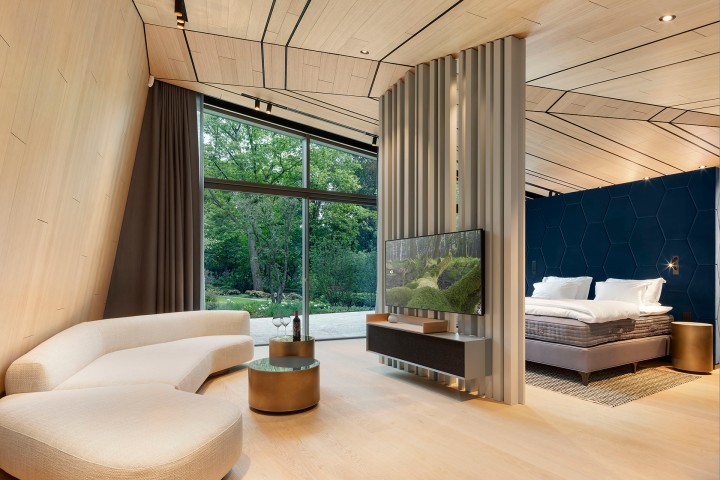 Modernes Hotelzimmer in hellen Beige- und Brauntönen, helles Holz an Wänden, Decke und Böden, cremefarbenes futuristisches Sofa, Flatscreen an einem Raumtrenner, Blick durch eine große Fenterfront in einen Garten