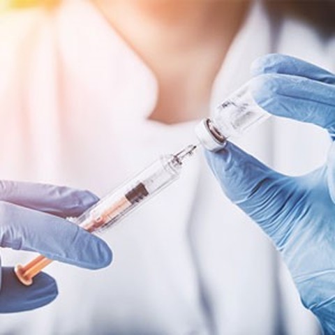 Quadratisches Bild, auf dem man die Hände einer Ärztin mit blauen OP-Handschuhen sieht, die eine Spritze mit einer Flüssigkeit aus einer Ampulle aufzieht