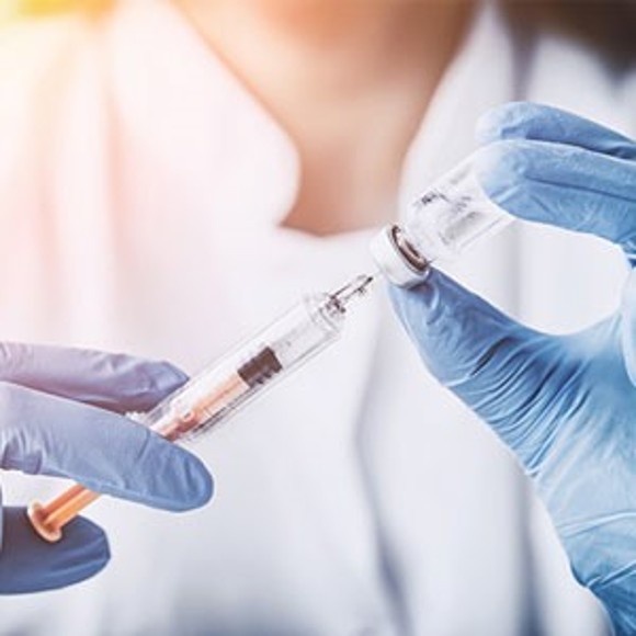 Quadratisches Bild, auf dem man die Hände einer Ärztin mit blauen OP-Handschuhen sieht, die eine Spritze mit einer Flüssigkeit aus einer Ampulle aufzieht