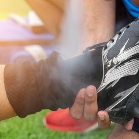 Quadratisches Bild, auf dem man sieht, wie die Hände eines Mannes einen Fuß mit schwarzem Fußballschuh stützen und etwas aufsprühen
