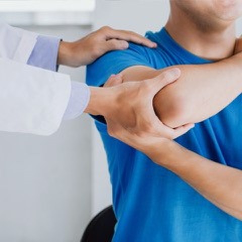 Quadratisches Bild, auf dem man die Hände eines Arztes in weißem Kittel sieht, die den Ellbogen und Arm eines männlichen Patienten im blauen Shirt behandeln.
