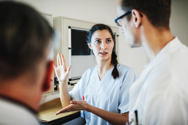 Es sind zwei Ärzte von hinten zu sehen, während eine Krankenschwester in blauer Arbeitskleidung an einem Tresen lehnt und beiden etwas erklärt, während sie mit den Händen gestikuliert