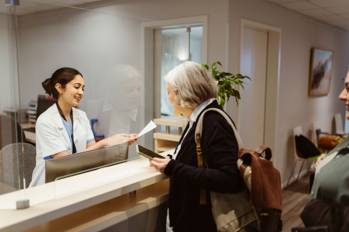 Ältere Patientin steht mit einem Handy in der Hand am Empfangstresen einer Arztpraxis und eine junge Arzthelferin kümmert sich lächelnd um sie