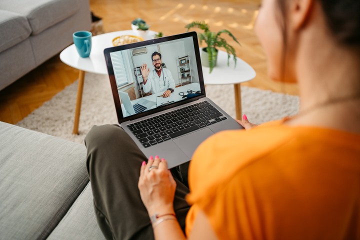 Junge Frau mit orangefarbenem Oberteil - unscharf im Vordergrund des Bildes - sitzt im Schneidersitz mit einem laptop auf dem Schoß auf einem Sofa und erhält per Videocall eine Beratung eines Arztes