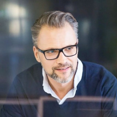 Lächelnder Mann mittleren Alters mit gepflegtem grauen Bart und nach hinten gegelten Haaren, schwarzer Kunststoffbrille, blauem Pullover und weißem Hemd darunter