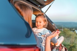 Kleines, blondes Mädchen im blau-weiß geringeltem Pullovert sitzt in der Abendsonne im Kofferraum eines Autos und streckt seiner Mutter die Zunge heraus.