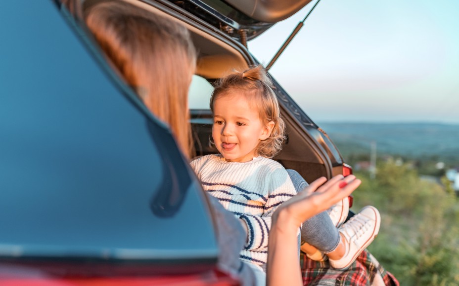 Kleines, blondes Mädchen im blau-weiß geringeltem Pullovert sitzt in der Abendsonne im Kofferraum eines Autos und streckt seiner Mutter die Zunge heraus.