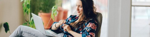 Junge Frau mit langen, dunklen Haaren in einer geblümten Bluse sitzt entspannt mit einer blauen Kaffeetasse in der Hand und dem Laptop auf dem Schoß, im Hintergrund terrakottafarbene Pflnztöpfe und eine Grünpflanze