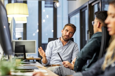 Ein Mann erklärt etwas während eines Gesprächs mit Kollegen in einem modernen Büro. Die Szene zeigt Austausch und professionelle Zusammenarbeit.
